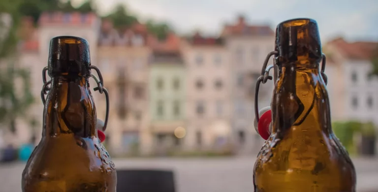 Zwei Glasbierflaschen mit geöffneten Verschlüssen vor den malerischen Häusern von Ljubljana an einem Fluss am Abend.